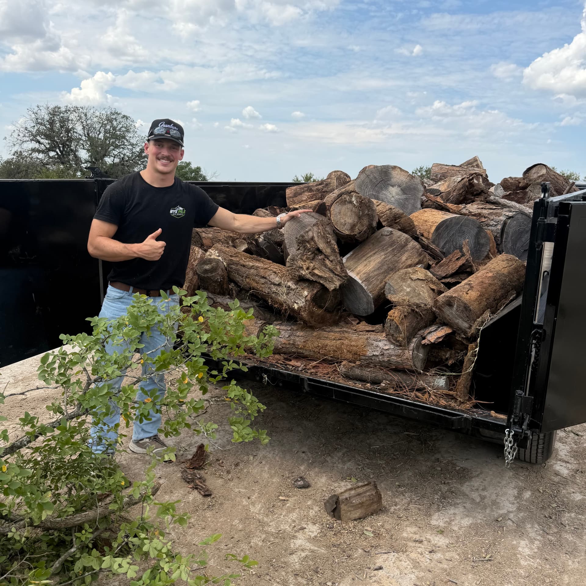 Bye Pile Junk Removal owner giving a thumbs-up beside a dump trailer loaded with logs